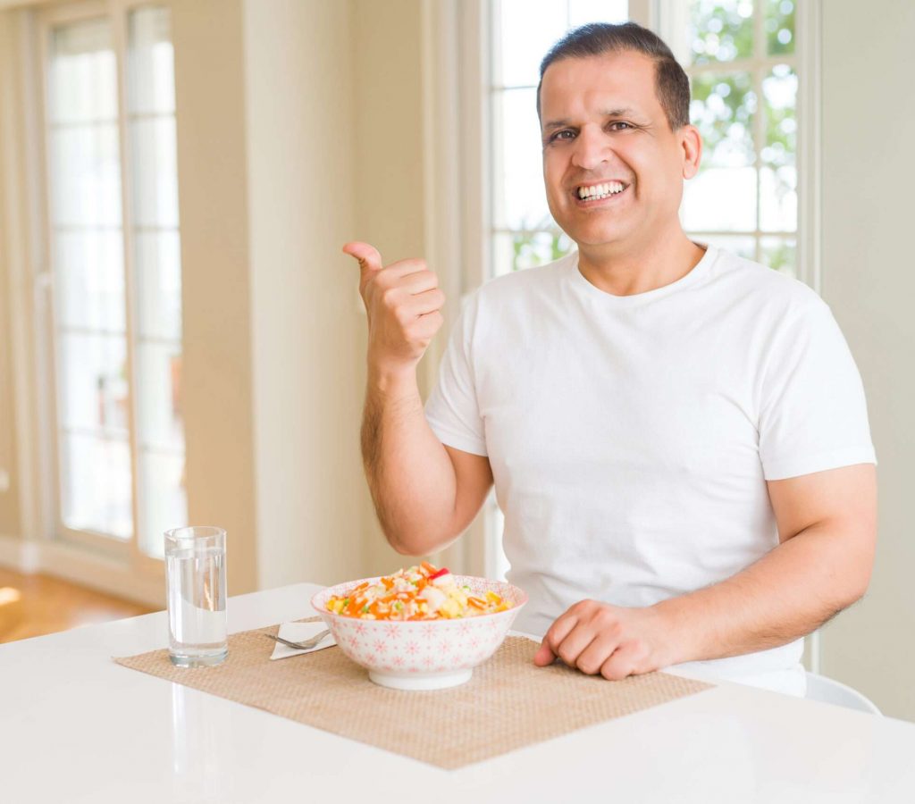 Middle age man eating rice at home smiling with happy face looking and pointing to the side with thumb up.